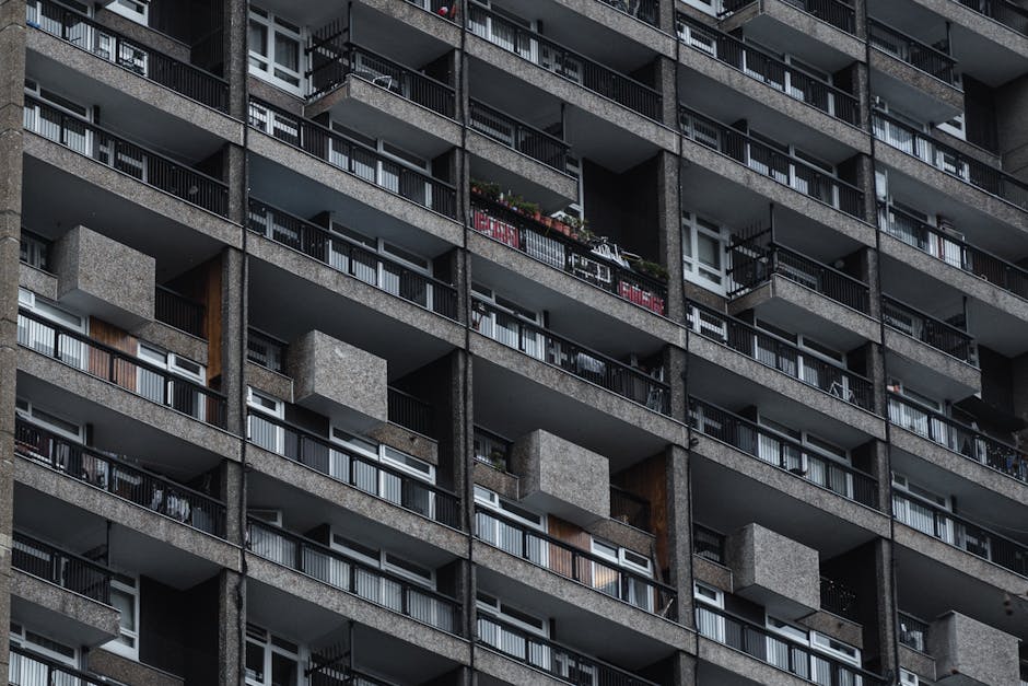 A close-up view of the exterior façade of a high-rise residential building made of concrete with multiple balcony levels. Each balcony features metal railings, some with visible flowers and outdoor f