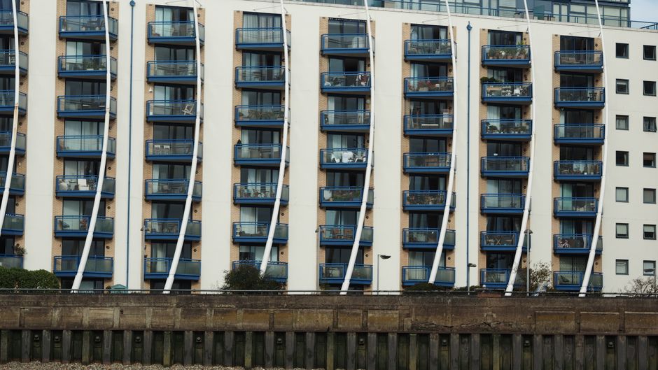 A close-up view of the exterior façade of a high-rise residential building made of concrete with multiple balcony levels. Each balcony features metal railings, some with visible flowers and outdoor f